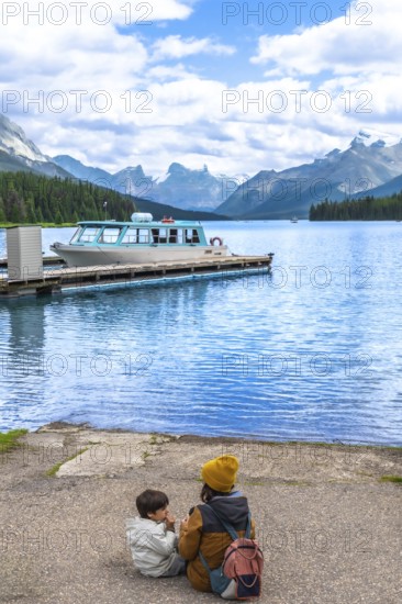 Mother and son sitting on maligne lake's shore, admiring the turquoise water and a tour boat at the
