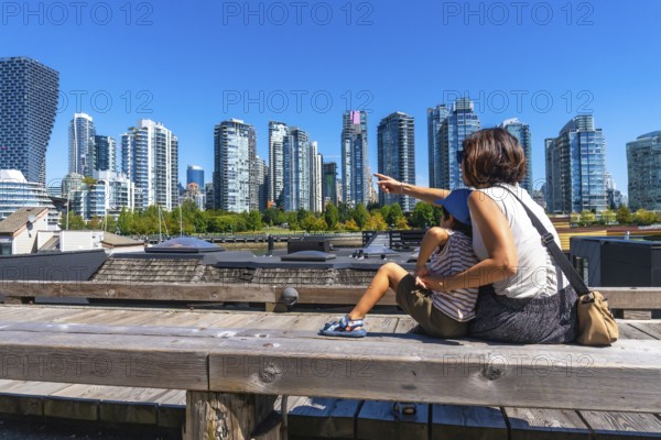 Tourist mother pointing at the skyline of vancouver while sitting with her son on a wooden bench at