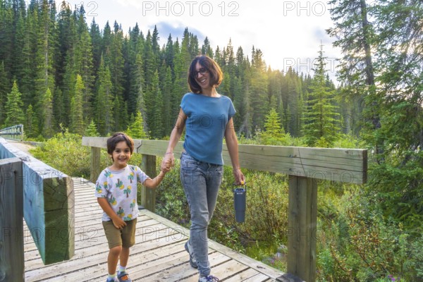 Mother and son holding hands, smiling and walking on a wooden boardwalk surrounded by lush green
