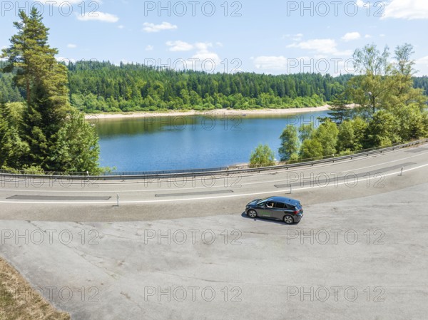 A black car drives along a lake, surrounded by a forest, in clear skies, deer e-car sharing, Cupra
