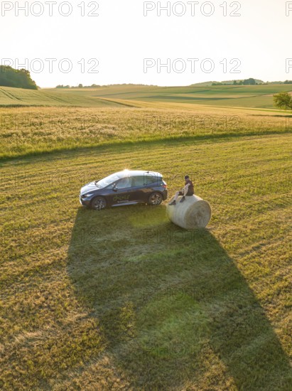 Car with person on a hay bale in a wide field at sunset, deer e-car sharing, Cupra electric car,