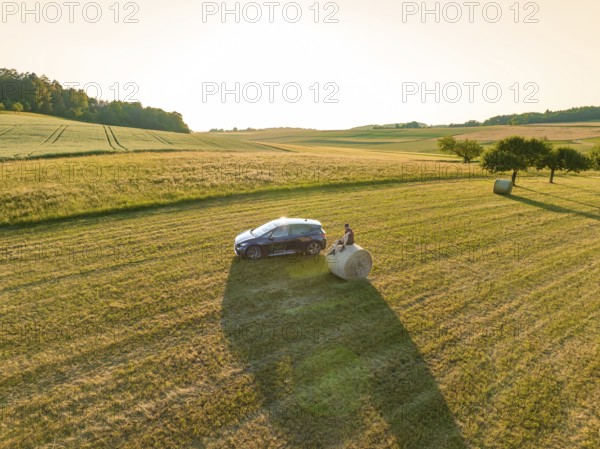 Car next to a hay bale in the middle of a vast golden field under the evening sun, deer e-car