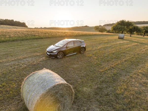 A car is parked in a field with hay bales at sunset, surrounded by meadows, deer e-car sharing,