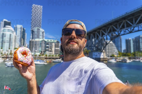 Tourist taking a selfie while holding a donut with burrard bridge and the cityscape of vancouver in