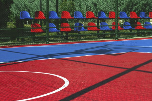 A vibrant basketball court features a mix of red and blue colors. Empty seats line the side,
