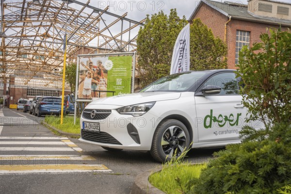 An Opel with car sharing logo stands in an urban environment with billboard and vegetation, Deer