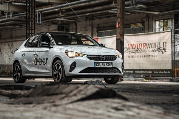 A white car is parked in front of a Motorworld banner in an industrial hall, Deer E-Car Sharing,