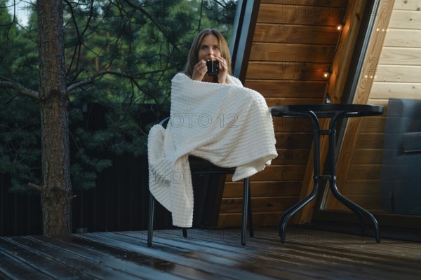 A woman relaxes on a chair while wrapped in a blanket. She holds a drink and enjoys fresh air after