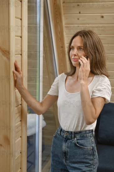 A woman stands near a wooden wall in a modern room while talking on her phone. She appears focused