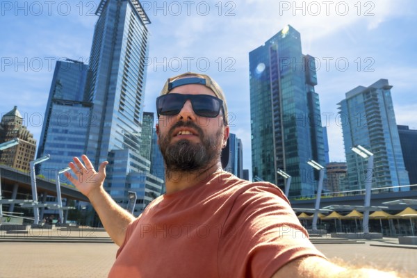 Bearded tourist wearing sunglasses and baseball cap takes selfie with outstretched arm in front of