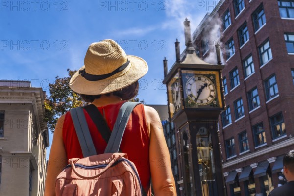 Female tourist wearing straw hat and backpack looking at the famous steam clock in gastown historic