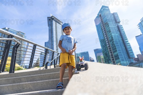 Young boy standing on steps playing with a toy car, with vancouver's modern buildings and clear
