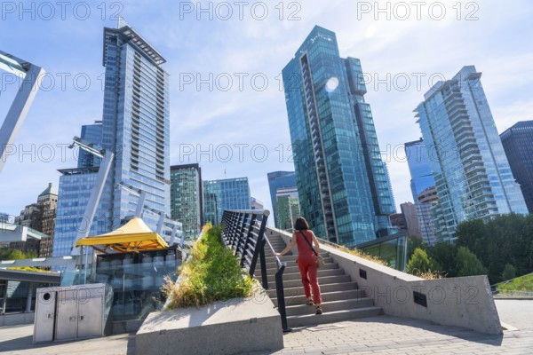 Woman is climbing the stairs of a pedestrian overpass with modern skyscrapers and a clear blue sky