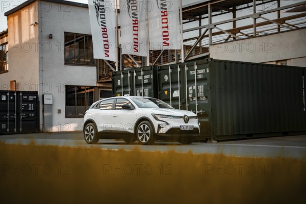 White modern SUV in front of an industrial building with containers and banners, Deer E-Carsharing,