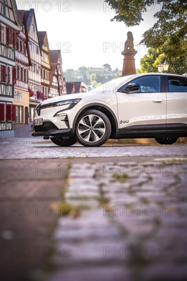 White car on cobblestone road surrounded by half-timbered houses and trees, Deer E-Carsharing,