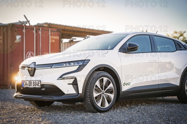 A white electric car stands on a gravel field in front of a container at dusk, Deer E-Carsharing,