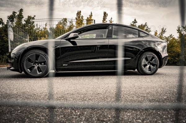 Side view of a black car in front of a fence surrounded by autumn leaves, Deer E- car sharing,