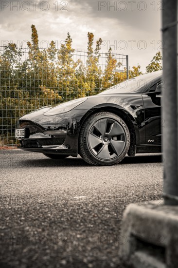 Black car on asphalt in front of a fence surrounded by trees and clouds, Deer E-Carsharing, Tesla