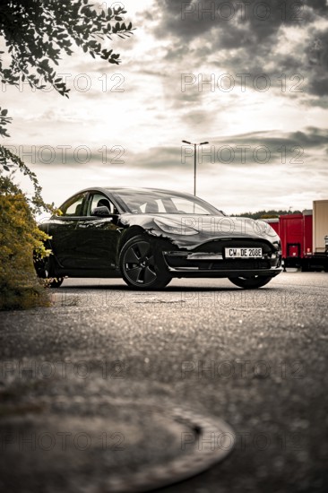 Black car in a parking lot under cloudy sky with trees nearby, Deer E- car sharing, Tesla Model 3,