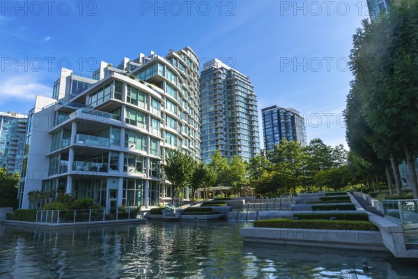 Modern glass and concrete buildings stand tall, reflecting in the calm water of a city park,