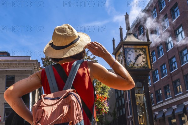 Young woman with backpack and straw hat looking at the famous steam clock in gastown, vancouver,