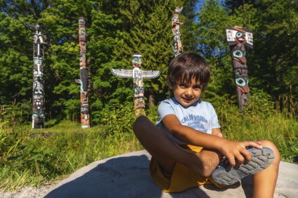 Young boy sitting on a rock, tying his shoe in front of vibrant totem poles at brockton point in