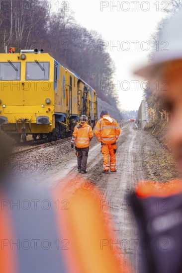 Two workers in orange on a railroad construction site next to a large yellow vehicle, tamping