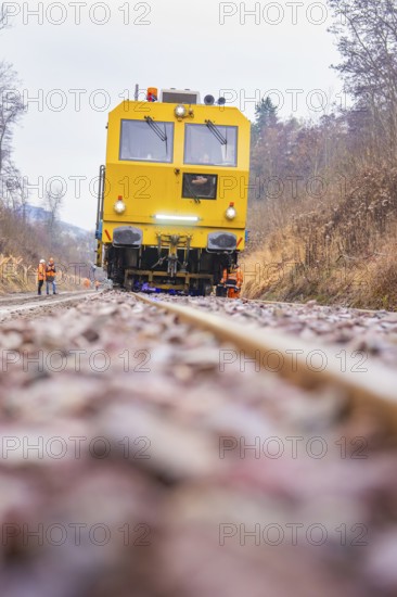 Yellow train on dirt road through forest with workers in the background, stuffing machine on