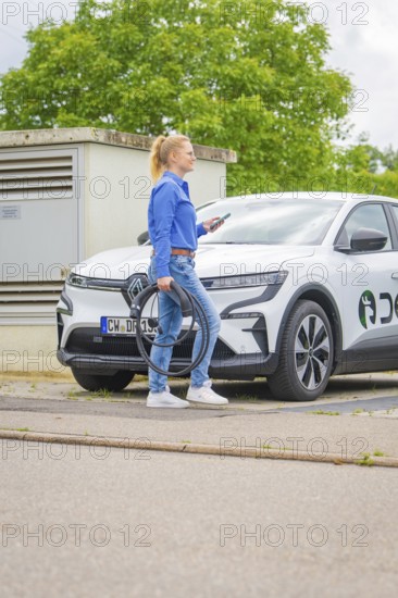 Woman standing on the street with a white electric car with charging cable, Deer E-Carsharing,