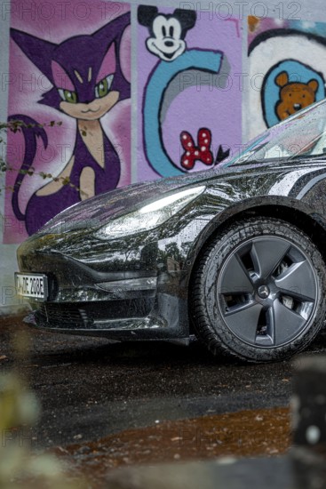 Close-up view of a black car in front of colorful graffiti art on a wet road, Deer e-car sharing,