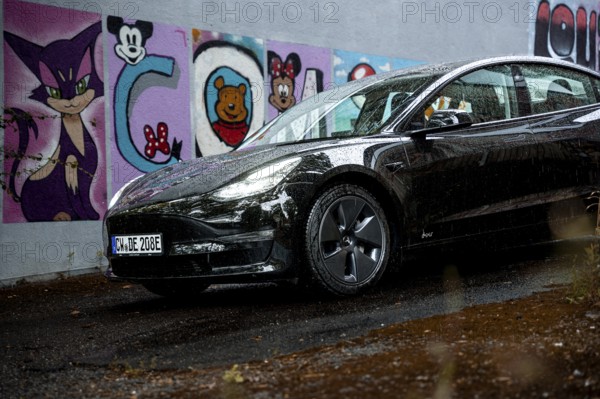 Black car in front of a rain-covered road and colorful graffiti wall, Deer E-Carsharing, Tesla
