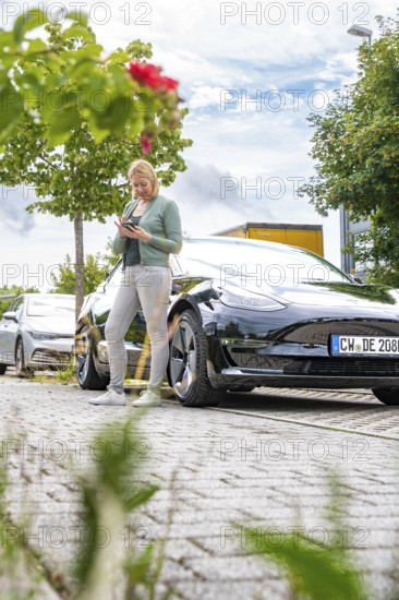 Woman using a tablet next to a parked black car outdoors, Deer E- car sharing, Tesla Model 3, Calw,