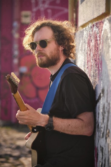 Hipster street musician in black playing electric guitar in the street on sunset leaning on a wall.