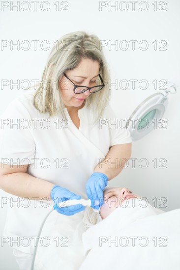 Cosmetologist in blue gloves delivers a hydrafacial cleansing treatment to a client, using blue