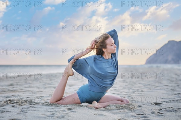 A woman practices Eka Pada Rajakapotasana on the sand during sunset, focusing on her yoga pose with