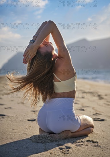 Woman performs Vajrasana with backbend on sandy beach as the sun sets over the ocean. She kneels on