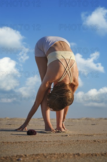 A woman performs Uttanasana on a sandy beach during sunset while clouds fill the sky. She leans