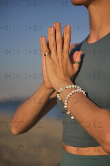 A woman practices yoga by holding hands in Namaskar Mudra at the beach as the sun sets in the