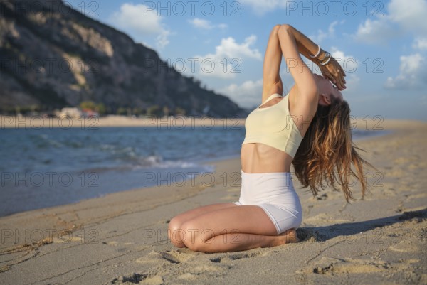 A woman kneels on a sandy beach, performing Vajrasana with a deep backbend and arms raised overhead