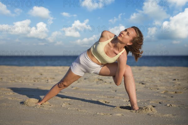 A woman practices Baddha Trikonasana on a sandy beach with wide legs and arms bound. The sun sets