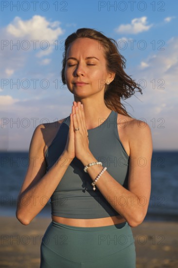 A woman practices yoga on the beach at sunset, holding hands in Namaskar Mudra as she meditates