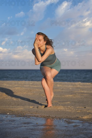 A woman practices standing Garudasana on a beach with one leg wrapped around the other at sunset by