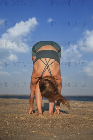 A woman does Uttanasana on a sandy beach during sunset, focusing on her breath and stretching her