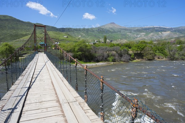 View of a swinging suspension bridge spanning a river, with green hills and a mountain in the