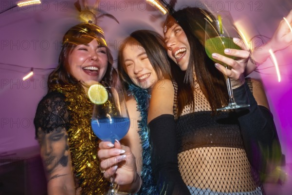 Three happy young adult women laughing while holding colorful cocktails, celebrating together with
