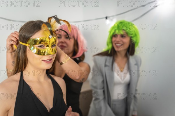 Friends preparing for a celebration, with one woman helping another put on a gold masquerade mask