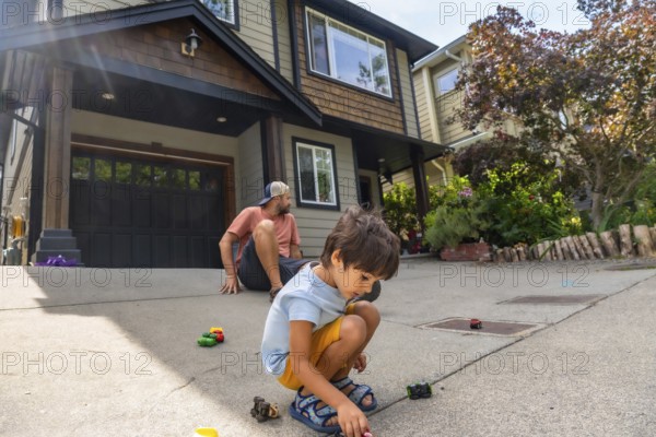 Young boy plays with toy cars on the driveway of a modern home while his father watches from
