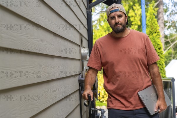 Maintenance worker walking alongside a residential building, carrying a laptop and various tools,