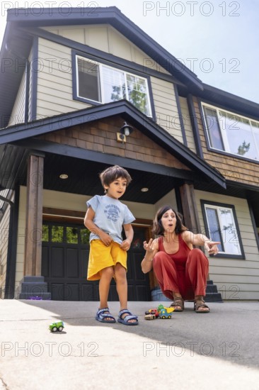 Mother and son are crouching down, playing with toy cars on the driveway in front of their house,