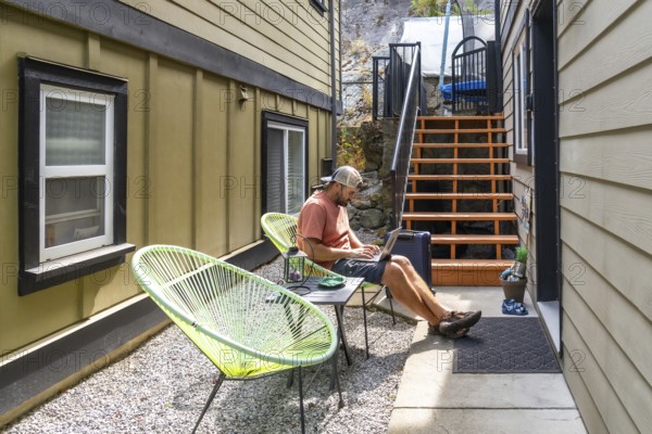 Young man working remotely on a laptop, lounging comfortably in a patio chair in his backyard,